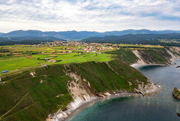 Aerial view of a coastal village surrounded by green fields, cliffs, and a calm blue sea with mountains in the distance under a cloudy sky