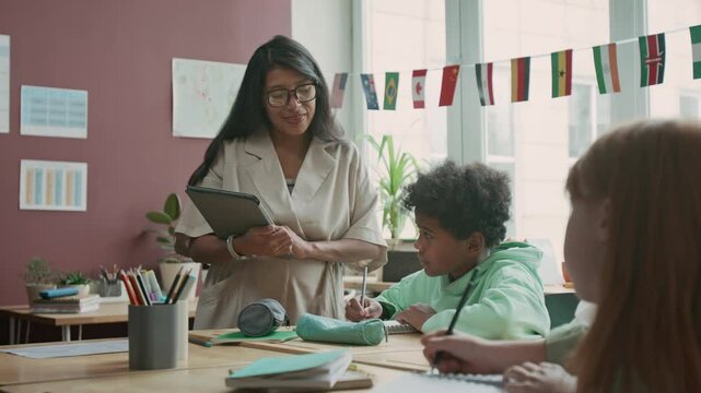 Medium shot of smiling Hispanic female teacher standing in classroom at foreign language lesson, watching African American boy and Caucasian girl writing dictation in notebooks
