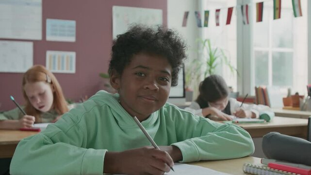 Medium portrait of 10-year-old immigrant African American boy with curly hair sitting at desk in English language school with diverse classmates, writing test on sheet, looking at camera and smiling