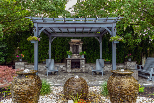 Large gray pergola on pavers with stone fireplace and waterfall urns for backyard oasis landscape design