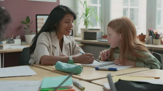 Medium zoom-in shot of multiethnic boy and girl writing exercise in copybooks during English language lesson, while Hispanic female teacher is explaining rules to red-haired Caucasian schoolgirl