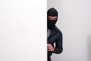A criminal wearing balaclava peeking from behind white wall