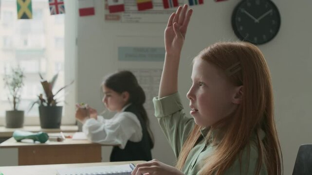 Medium close-up side shot of 10-year-old Caucasian girl with long red hair sitting at desk in classroom during foreign language lesson, writing in copybook, then raising hand to answer question