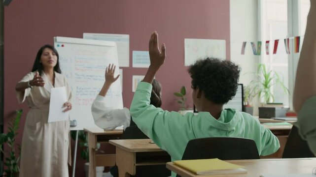 Medium full shot of young female teacher explaining new topic at foreign language lesson at school for migrant children, asking questions to diverse class, all children raising hands
