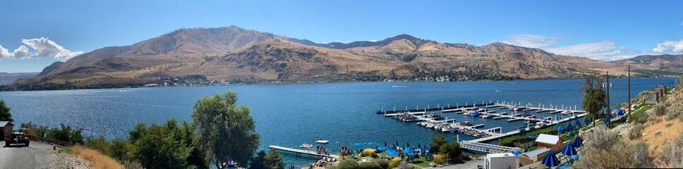 A panoramic view of Lake Chelan in Washington state showing a marina in the right foreground and the arid hills in the background on a beautiful summer day_08252024_0003.