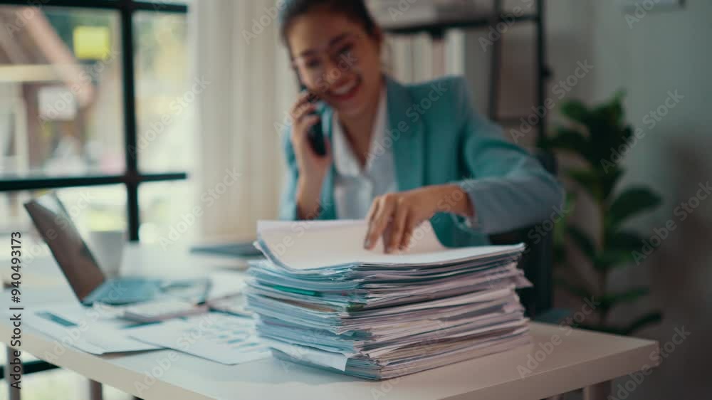 Poster businesswoman searching through a large stack of paperwork while smiling and talking on her cell pho