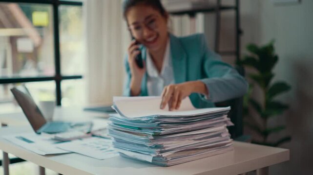 Businesswoman searching through a large stack of paperwork while smiling and talking on her cell phone. A busy office environment highlights her multitasking and professional demeanor