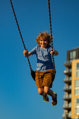 Kid swinging on chain swing on city kids playground. Swing ride. Cute child having fun on a swing on summer sky background. Blonde little boy swings at kid playground. Child swinging high.