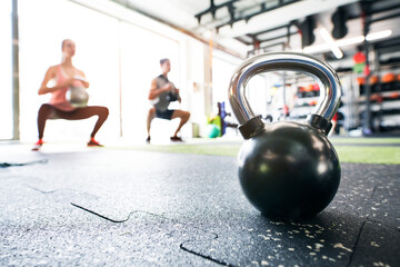 Kettlebell on floor in gym, people doing kettlebell swing behind. Gym equipment for strength training.