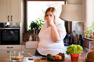 Overweight woman in the kitchen, drinking cup of fresh tap water. Choosing healthy diet, making vegetable salad.