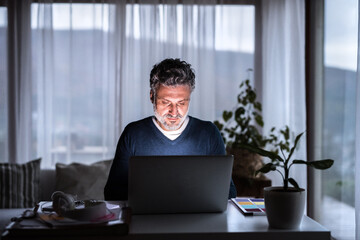 Handsome man working from home office at evening, using laptop. Screen glow illuminates man's face....