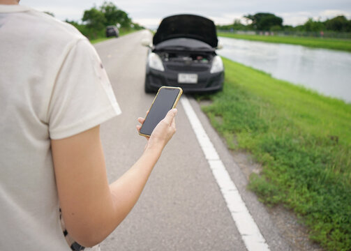 Woman's hand using smartphone to call auto mechanic with broken car on the roadside. A woman is holding a mobile phone and calling for help with a broken down car on the road. - Powered by Adobe