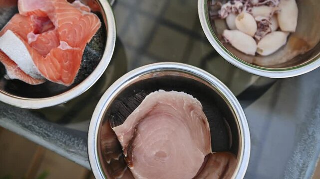 Fresh seafood ingredients prepared for a barbecue, showcasing a variety of raw fish and squid rings in stainless steel bowls on a kitchen counter