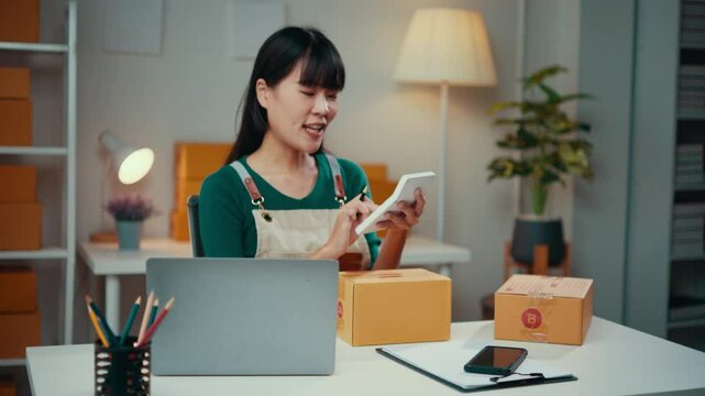 Young asian woman working as an sme entrepreneur, managing a laptop while packaging customer orders in boxes from her home office. Focused on online selling and customer service