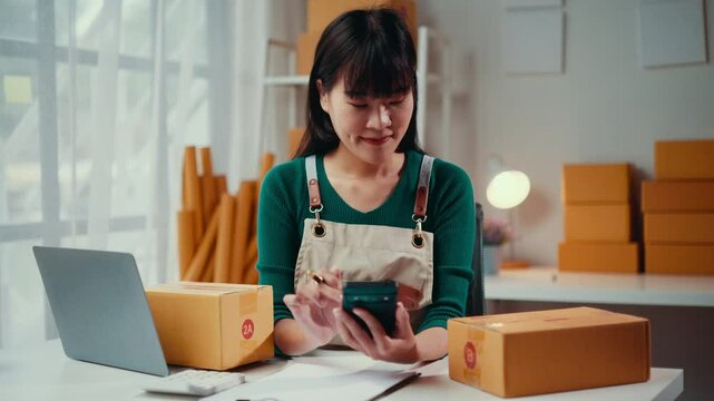 Young asian woman entrepreneur using calculator and smartphone checking customer order packing products into boxes, preparing it for delivery