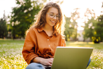 A young woman enjoying a sunny afternoon working on her laptop in a park setting surrounded by greenery and urban views. Freelancer, Online education.