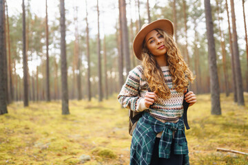 Young woman in a forest wearing a hat and backpack enjoys a peaceful day hiking among tall trees during autumn. Forest baths. Active lifestyle