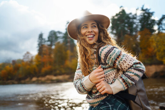 A young traveler woman enjoying a sunny autumn afternoon by the riverside with a backpack and a cheerful smile. The concept of hiking, travel, vacation.