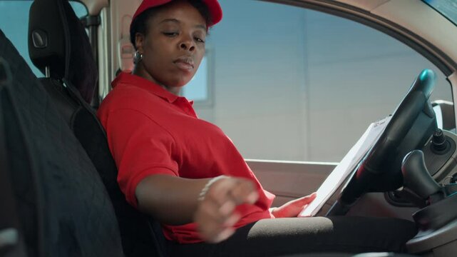Side view of female African American courier in red cap and uniform checking delivery forms while sitting in van