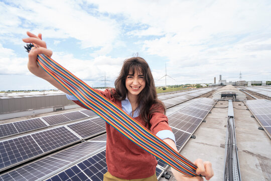 Smiling businesswoman holding multi colored wire strap and standing on rooftop with solar panels