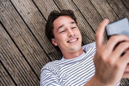 Smiling young man surfing net through smart phone lying down on wooden patio