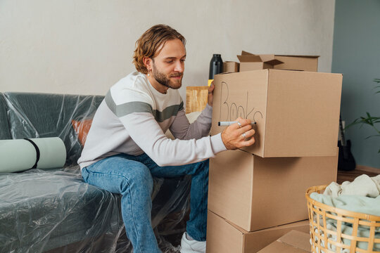 Young man writing on cardboard box sitting in living room at home