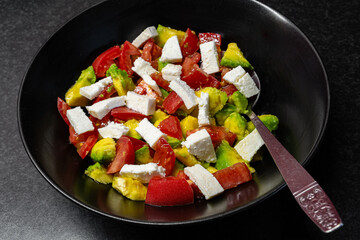 Fresh red tomato, avocado and bryndza or feta salad with olive oil in black dish with a spoon close-up view from top, healthy eating.