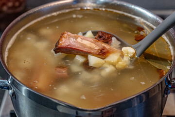 Soup on meat clear broth of smoked pork beef ribs with potatoes, cauliflower, bay leaf and salt in a stainless steel pot close-up with a plastic ladle.