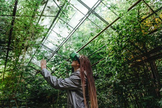 Woman gesturing and watching trees through VR glasses in greenhouse