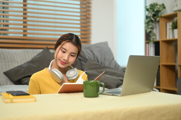 Smiling young asian girl sitting in living room using laptop and writing in a notebook