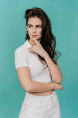 Fototapeta premium thoughtful young woman in a white cropped polo shirt and white pants, with her hand on her chin, looking to the side against a turquoise background