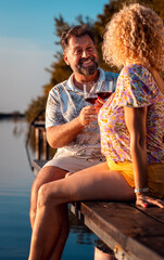 Smiling adult couple sitting by the lake on a pier drinking wine during sunset.