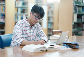 Obraz premium Young Asian male sitting inside a library alone doing research. Man working on a project. Young man doing research for a case.
