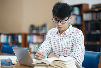 Young Asian male sitting inside a library alone doing research. Man working on a project. Young man doing research for a case.