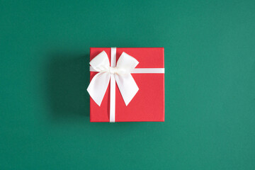 Red square gift box, present with white satin ribbon with bow on green background. Christmas. Flat lay, top view, copy space