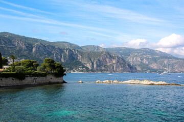 Coastal landscape of Saint-Jean-Cap-Ferrat, French Riviera, showcasing clear Mediterranean waters, a stone coastline, and rugged mountains in the background. 