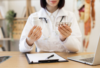 Doctor holding bottle of tablets and blister pack of pills in medical office. Concept of healthcare professional prescribing medicine. Shot in clinical setting with laptop and clipboard on table.