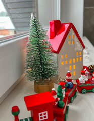 on the windowsill there is a red wooden retro train, a green Christmas tree, a ceramic house decorated with a garland with a star. A Christmas card. Vertical photo