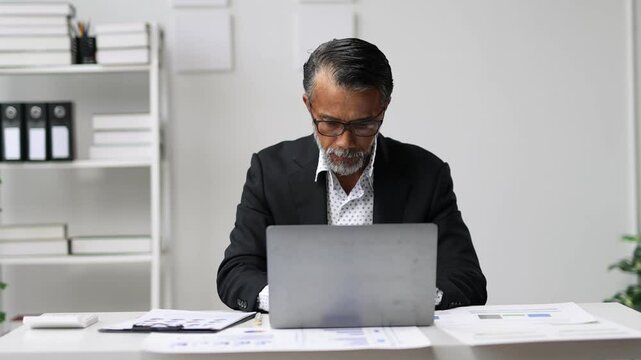 Focused businessman analyzing reports and working on a laptop in a modern office setting. Professional and productive, he is immersed in his work, managing documents and making notes.