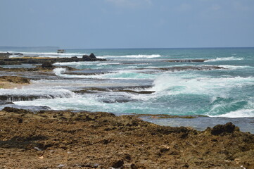 A scenic shot of waves breaking and flowing over rocky formations along a sandy beach, under a bright blue sky. The image captures the natural beauty and power of the ocean