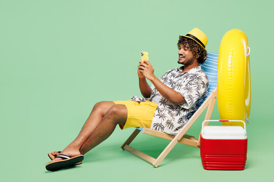 Full body side view young happy Indian man wear beach shirt hat rest near hotel pool sit in deckchair hold use mobile cell phone isolated on plain green color background. Summer sea sun tan concept.