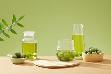 On green background, glass bottles of olive oil displayed with bowls of fresh olives and green leaves. Front view, empty space for presentation product
