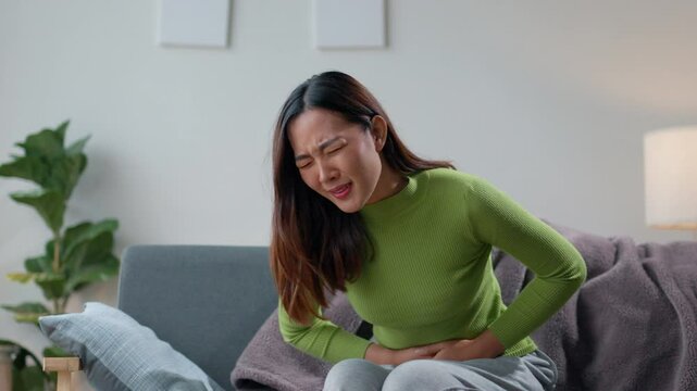 Young woman is sitting on her couch, hunched over in pain as she clutches her stomach. Her face is contorted in discomfort