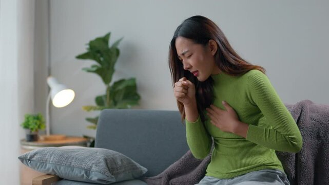 Young woman sits on a sofa in her living room, coughing and holding her chest. Signs of illness are evident as she struggles with discomfort and worries about her health