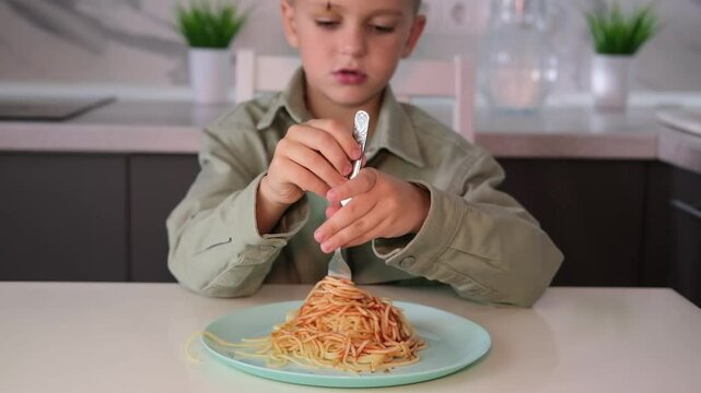 Boy eats spaghetti with appetite. Funny child gets dirty eating pasta, wraps it on fork from large plate at home. Baby food concept.