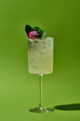 A refreshing summer cocktail served in a tall glass with a thick base. The drink is garnished with a sprig of mint and a pink flower, sitting on a bed of crushed ice.