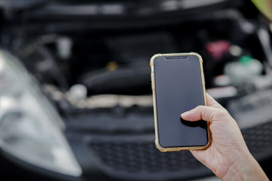 Close-up of woman's hand using smartphone to call auto mechanic with broken car on the roadside. Female hand holding a mobile phone with the background of a broken car