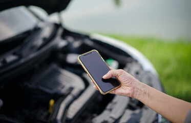 A woman's hand is using a smartphone to contact a mechanic with a broken car on the side of the...