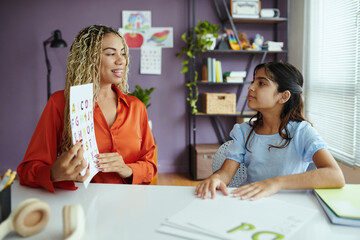 Teacher holding up alphabet chart while sitting across young girl in classroom environment with educational decorations and supplies visible in the background