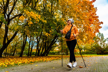 Beautiful middle-aged blonde woman dressed sporty walking with poles on alley full of yellow fallen leaves in park on autumn day. Front view. Autumnal training of Nordic walking.